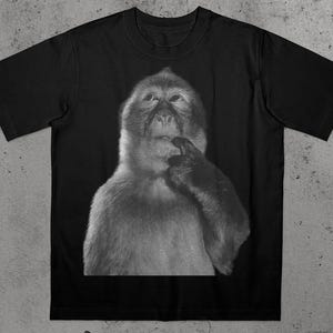 Peut inclure: T-shirt noir avec une image en niveaux de gris d'un singe dans une pose contemplative, le doigt sur les lèvres. La fourrure du singe est détaillée et le t-shirt a un col rond classique.