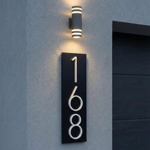 May include: A contemporary outdoor wall light and a black address plaque displaying the numbers 168. The light fitting is cylindrical with white horizontal stripes. The address plaque is rectangular with white numbers.