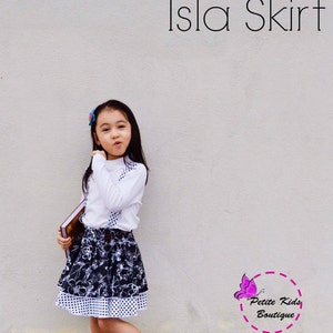 Puede incluir: Una niña con una camisa blanca de manga larga y una falda floral en blanco y negro con dobladillo de lunares. La falda está etiquetada como "Isla Skirt". Lleva zapatos blancos y sostiene un libro. El logotipo de "Petite Kids Boutique" es visible.