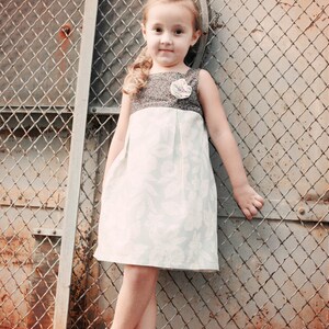 May include: A young girl wearing a sleeveless dress with a gray bodice and a light blue floral skirt. The dress has a flower detail on the shoulder. She is wearing white flip-flops and standing in front of a chain-link fence.