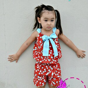 May include: A red and white polka dot romper with a blue bow. The romper has a gathered waist and shorts. The text "Johannah Romper" is at the top of the image. The text "Petite Kids Boutique" is at the bottom of the image.