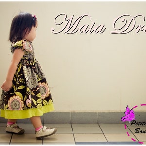 Può includere: Una bambina indossa un vestito floreale marrone e verde con una sottogonna gialla. Il vestito ha un collo a volant e maniche corte. La bambina è in piedi su un pavimento piastrellato e guarda a destra. Il testo "Maia Dress" è scritto in corsivo sopra la testa della bambina. Il testo "Petite Kids Boutique" è scritto in un cerchio con un disegno di farfalla sotto la bambina.