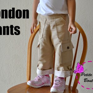 May include: A child wearing a pair of beige cargo pants with a white shirt. The pants have a white trim at the bottom and a button on the pocket. The text "London Pants" is visible at the top of the image.