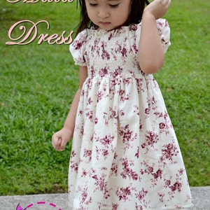 May include: A white dress with a floral print and a white smocked bodice. The dress has a white underskirt and is worn by a young girl with a white flower headband. The text "Maia Dress" is superimposed on the image.