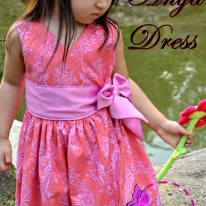 May include: A young girl wearing a pink and coral floral print dress with a pink sash and bow. The dress has a V-neck and is sleeveless. The girl is holding a green and red flower. The text "Anya Dress" and "Petite Kids Boutique" is visible in the image.