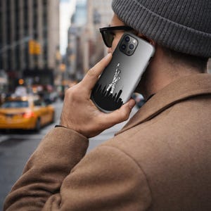 May include: A smartphone case featuring a black and white graphic of the New York City skyline, including the Statue of Liberty. The case has a gradient gray background and is held by a person wearing a gray beanie and brown coat.