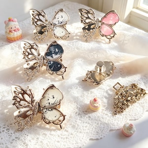 May include: A collection of gold-toned butterfly hair clips featuring intricate cut-out wings, clear rhinestones, and decorative inserts in white, pink, and dark blue with speckled patterns, accompanied by small miniature cupcake-shaped figurines.