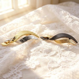 May include: Two hair barrettes with a curved, wave-like design. The left barrette features a gold-toned top layer with a textured finish over a silver-toned base. The right barrette has a matte black top layer over a gold-toned base.