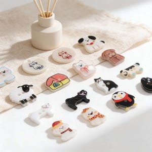May include: A collection of decorative buttons in various shapes, including animals, a house, and a snowman. The buttons are in a variety of colors, including white, black, brown, and yellow. Some buttons have painted details. A beige diffuser with wooden sticks is in the background.
