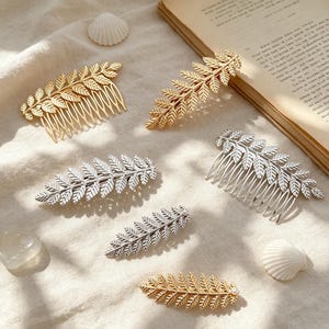 Op de afbeelding: Een collectie haaraccessoires in goud en zilver met bladmotieven. De set bevat haarkammen en haarspeldjes met gedetailleerde varenachtige bladvormen, sommige versierd met kleine witte parels, gerangschikt op een textiel ondergrond met textuur.