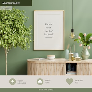 May include: A framed wall art print featuring the black text I'm not quiet. I just don't feel heard. centered on a white background. The print is displayed in a light wood frame hanging on a sage green wall above a wooden cabinet with a fluted design.