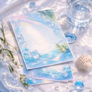 Könnte beinhalten: Briefpapier-Set mit einem Aquarelldesign einer Strandszene, einschließlich Regenbogen, Palmenblättern und Blasen. Das Set umfasst einen linierten Notizblock und kleinere Notizkarten. Klare Glasperlen und eine Muschel sind verstreut.