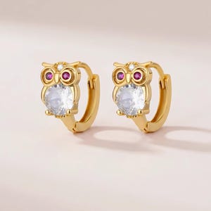 Könnte beinhalten: Ein Paar goldfarbene Creolen mit Eulenmotiv. Jede Eule hat zwei kleine runde rosa Steine als Augen, einen winzigen klaren Stein oben auf dem Kopf und einen großen, runden, klar facettierten Stein, der den Körper der Eule bildet.