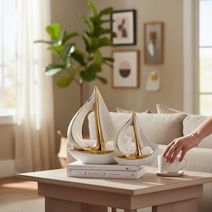 May include: Two white ceramic sailboat sculptures with gold accents on a wooden table. The sailboats have a modern design, with a curved sail and a gold base. The larger boat is approximately 30 cm tall.