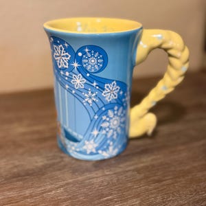 Może przedstawiać: Niebieski ceramiczny kubek z ż&oacute;łtym wnętrzem i uchwytem. Kubek ma wz&oacute;r płatka śniegu z białymi akcentami. Uchwyt ma kształt ż&oacute;łtego warkocza. Kubek stoi na drewnianej powierzchni.