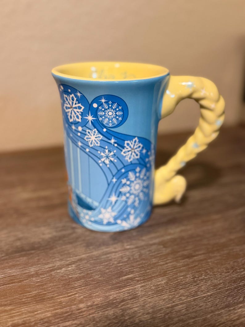Może przedstawiać: Niebieski ceramiczny kubek z ż&oacute;łtym wnętrzem i uchwytem. Kubek ma wz&oacute;r płatka śniegu z białymi akcentami. Uchwyt ma kształt ż&oacute;łtego warkocza. Kubek stoi na drewnianej powierzchni.