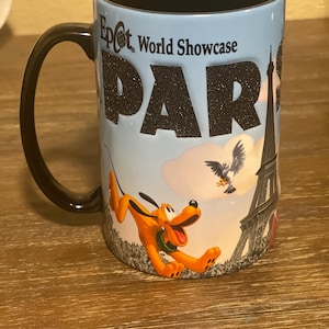 May include: A ceramic mug with a black handle and interior. The mug features the text "Epcot World Showcase PARIS" in black glitter lettering. Pluto is depicted running, with the Eiffel Tower and a bird in the background.