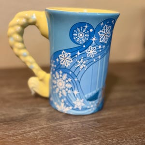 Może przedstawiać: Niebieski ceramiczny kubek z ż&oacute;łtym uchwytem i wnętrzem. Kubek ma zimowy motyw z białymi płatkami śniegu i wirującymi liniami. Uchwyt jest zaprojektowany tak, aby przypominał warkocz.