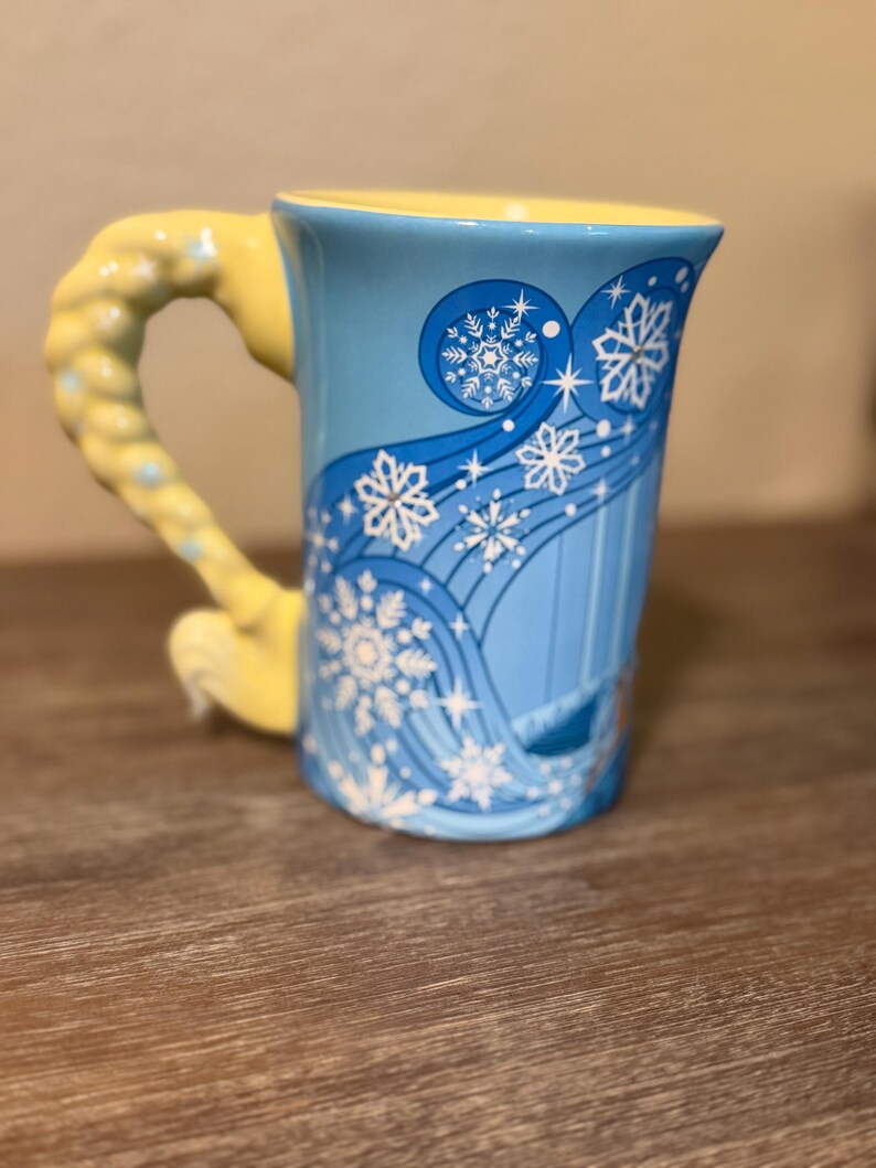 Może przedstawiać: Niebieski ceramiczny kubek z ż&oacute;łtym uchwytem i wnętrzem. Kubek ma zimowy motyw z białymi płatkami śniegu i wirującymi liniami. Uchwyt jest zaprojektowany tak, aby przypominał warkocz.