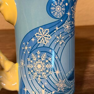 Może przedstawiać: Jasnoniebieski ceramiczny kubek z ż&oacute;łtym uchwytem. Kubek ma zimowy motyw z białymi płatkami śniegu i wirującymi niebieskimi liniami. Projekt zawiera stylizowaną sylwetkę zamku.