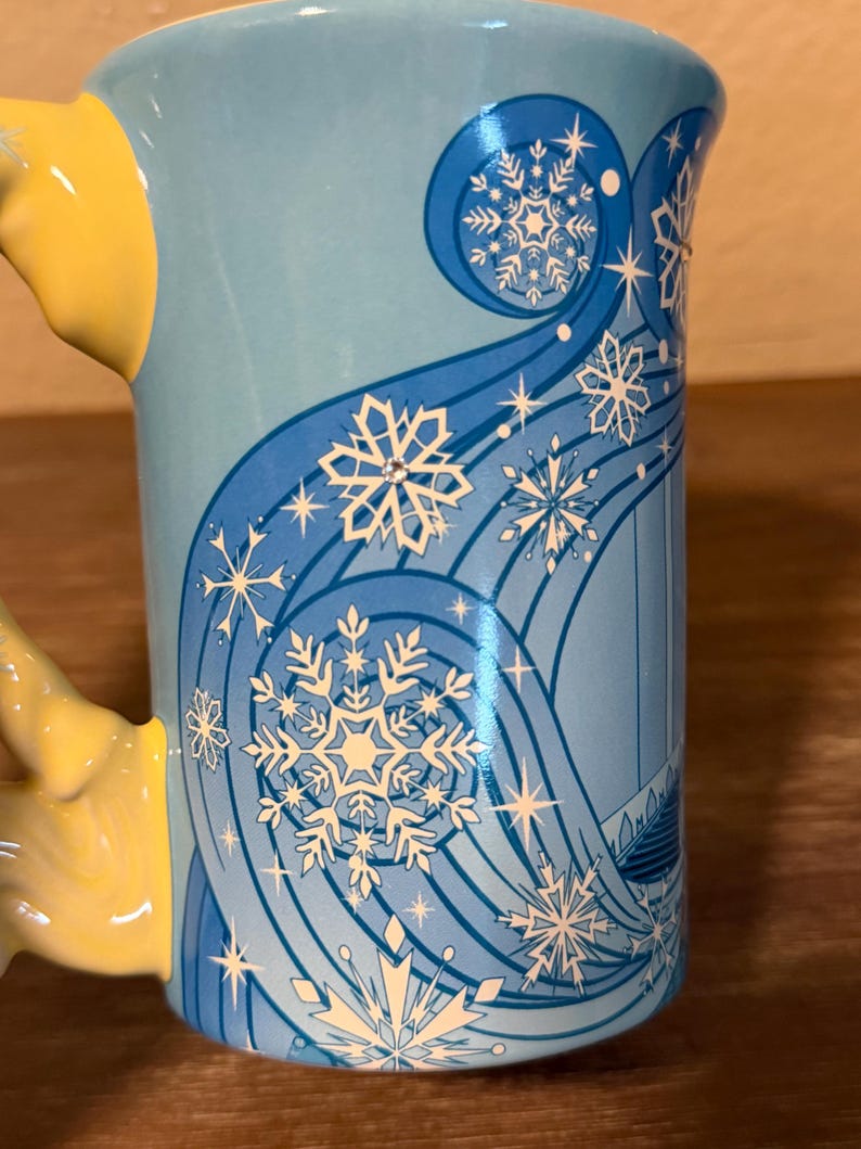 Może przedstawiać: Jasnoniebieski ceramiczny kubek z ż&oacute;łtym uchwytem. Kubek ma zimowy motyw z białymi płatkami śniegu i wirującymi niebieskimi liniami. Projekt zawiera stylizowaną sylwetkę zamku.