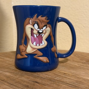 May include: A dark blue ceramic mug featuring a graphic of the Tasmanian Devil character with brown fur, a wide open mouth, and sharp white teeth. The mug has a curved handle on the right side and a smooth, glossy finish with a yellow interior rim.
