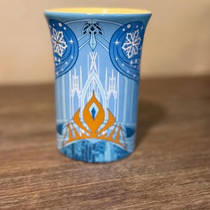 Może przedstawiać: Niebieski ceramiczny kubek z ż&oacute;łtym wnętrzem. Kubek ma wz&oacute;r z płatkami śniegu, zamkiem i pomarańczową koroną. Wz&oacute;r jest na jasnoniebieskim tle.