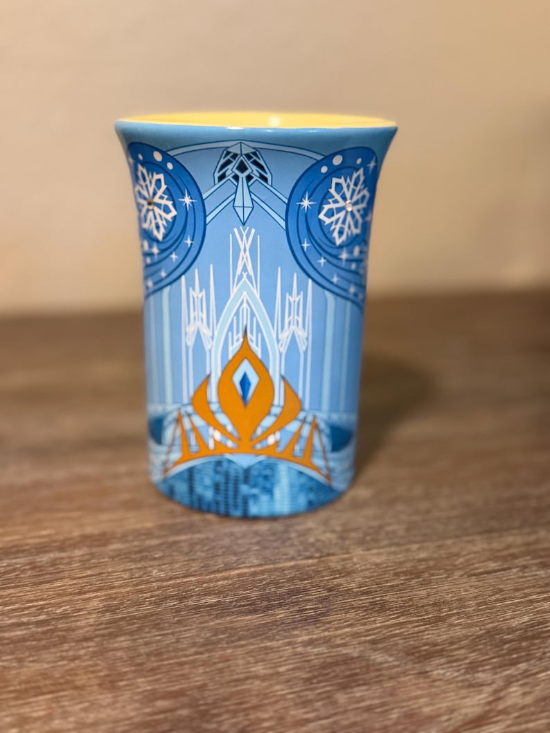 Może przedstawiać: Niebieski ceramiczny kubek z ż&oacute;łtym wnętrzem. Kubek ma wz&oacute;r z płatkami śniegu, zamkiem i pomarańczową koroną. Wz&oacute;r jest na jasnoniebieskim tle.