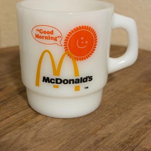Peut inclure: Un mug en verre opalin blanc arborant le logo McDonald's en jaune et noir, un soleil orange avec un visage souriant et une bulle contenant le texte "Good Morning" en orange. Le mug possède une anse robuste et une base à gradins.