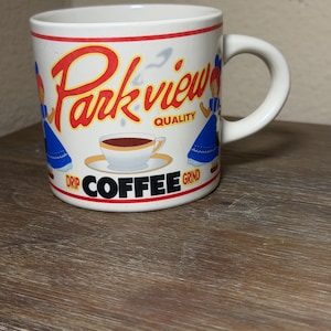 May include: White ceramic coffee mug with a curved handle. The mug features the words "Park View" in red script, along with illustrations of women in blue dresses and the words "Drip Coffee Grind".