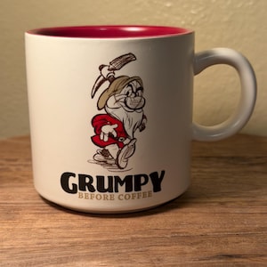 May include: A white ceramic mug with a red interior and a handle on the right side. The front features a sketch-style illustration of a bearded character wearing a hat and red tunic, with the text GRUMPY printed in black and BEFORE COFFEE in tan.