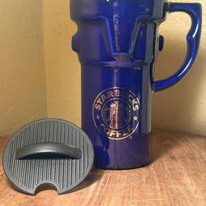 Può includere: Un mug alto in ceramica blu profondo con finitura metallica e manico abbinato. La parte anteriore presenta un logo circolare dorato Starbucks Coffee con una grafica a sirena. Un coperchio rotondo in plastica nera con texture a coste è posizionato davanti.