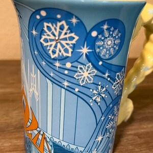 Może przedstawiać: Jasnoniebieski ceramiczny kubek z ż&oacute;łtym wnętrzem i uchwytem. Kubek ma zimowy motyw z płatkami śniegu i pomarańczowym napisem "FROZEN". Uchwyt to rzeźbiona figura w kolorze ż&oacute;łtym.