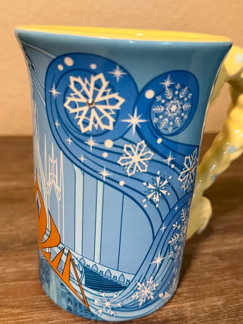 Może przedstawiać: Jasnoniebieski ceramiczny kubek z ż&oacute;łtym wnętrzem i uchwytem. Kubek ma zimowy motyw z płatkami śniegu i pomarańczowym napisem "FROZEN". Uchwyt to rzeźbiona figura w kolorze ż&oacute;łtym.