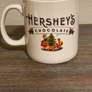May include: Cream-colored Hershey's Chocolate mug with a large handle. The mug features the Hershey's logo in brown script with a banner that reads "CHOCOLATE". A vintage illustration of children decorating a Christmas tree is below the logo.