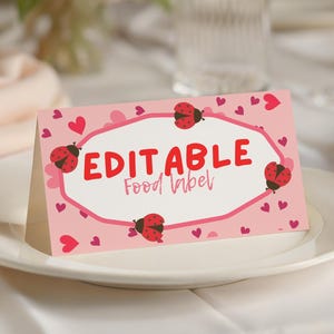 Peut inclure: Une étiquette alimentaire rose et pliable avec le mot "EDITABLE" en rouge, entourée de coccinelles et de cœurs. L'étiquette est posée sur une assiette blanche, avec un arrière-plan flou de fleurs et de verrerie.