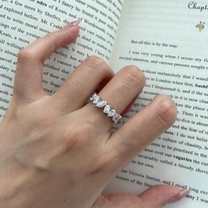 May include: A silver ring with multiple rectangular and marquise-cut clear gemstones. The ring is worn on a finger, with a book in the background.