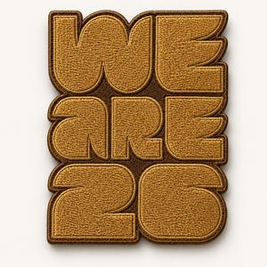 May include: A rectangular patch with the words "WE ARE 26" embroidered in a gold-colored thread on a brown background. The letters and numbers are bold and stylized, with a textured appearance.