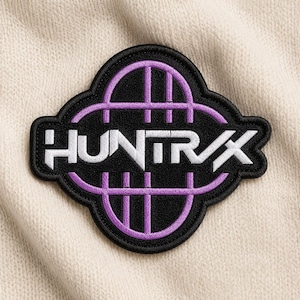 May include: A black and purple embroidered patch with the word "HUNTRX" in white. The patch has a unique shape with a purple grid design and a black border. The patch is on a cream-colored fabric.
