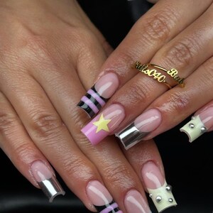 May include: A close-up of a hand with long, square-shaped acrylic nails. The nails feature a variety of designs, including French tips with silver accents, black and pink stripes, a pink nail with a yellow star, and blue polka dots. A gold ring with script is also visible.