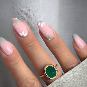 Pode incluir: Unhas ovais com base rosa, bolinhas brancas e desenhos florais brancos. Algumas unhas têm uma ponta verde clara. Um anel de ouro com uma grande pedra verde está no dedo anelar.