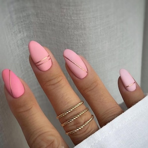 May include: Close-up of a hand with almond-shaped, matte pink nails. Each nail is adorned with a thin, metallic gold stripe. The ring finger features three gold, twisted band rings. The nails are a soft pink color.