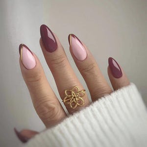 Puede incluir: Una mano con uñas almendradas pintadas en malva y rosa claro, algunas con borde dorado. Un anillo de flores de alambre dorado adorna el dedo anular. La mano lleva un suéter blanco. Manicura.