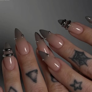 Puede incluir: Primer plano de uñas largas en forma de estilete con un diseño degradado nude a gris. Cada uña está adornada con pequeños tachuelas plateadas. Un dedo presenta un anillo plateado. Se ven tatuajes de formas geométricas y una telaraña.