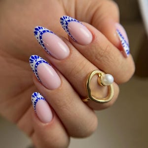 May include: Almond-shaped nails featuring a nude base with a blue and white intricate floral pattern along the tips in a French manicure style. A gold-toned ring with an irregular open band design and a single white pearl accent is worn on the finger.