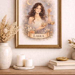 May include: A framed watercolor print featuring a Libra zodiac illustration. The artwork depicts a woman holding scales, surrounded by flowers and a crescent moon. The print is in a gold frame, displayed on a wooden surface with candles and decorative items.