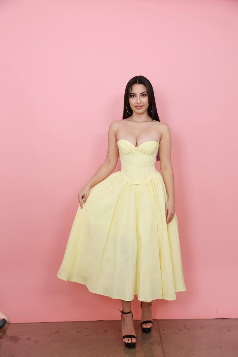 May include: A strapless, pale yellow dress with a fitted bodice and a full skirt. The dress features a sweetheart neckline and a bow detail. The model is wearing black heels. The background is a solid pink.