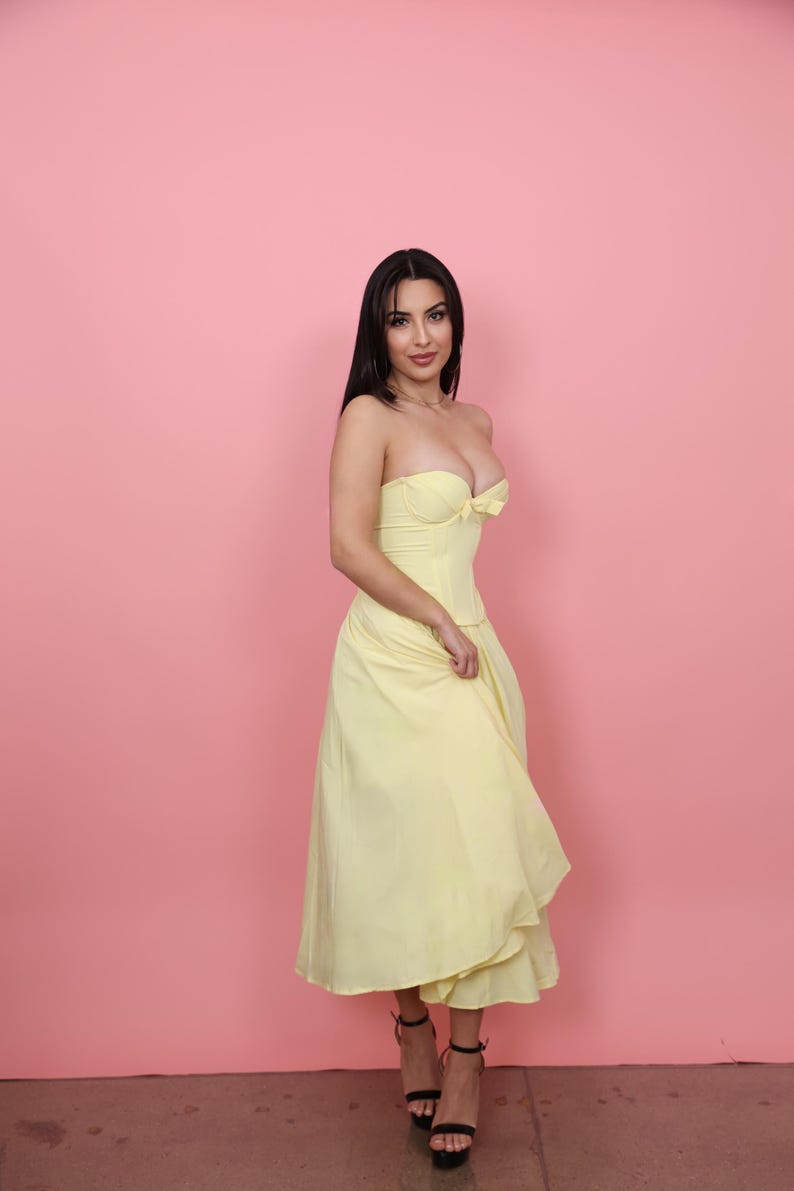 May include: A strapless, pale yellow dress with a fitted bodice and flowing skirt. The dress features a sweetheart neckline and a bow detail. The model is wearing black high-heeled sandals. The background is a solid pink.
