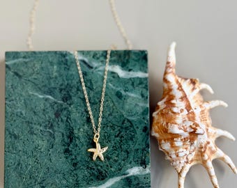 May include: A gold-colored starfish pendant necklace with a delicate chain, displayed on a green marble block. A seashell with brown and white tones is next to the block. The necklace is a jewelry item.