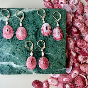 May include: Six pairs of earrings with pink and white shell pendants and gold-colored ear wires are displayed on a green marble surface. The earrings are accompanied by a pile of similar shells. Each earring has a small pearl bead.